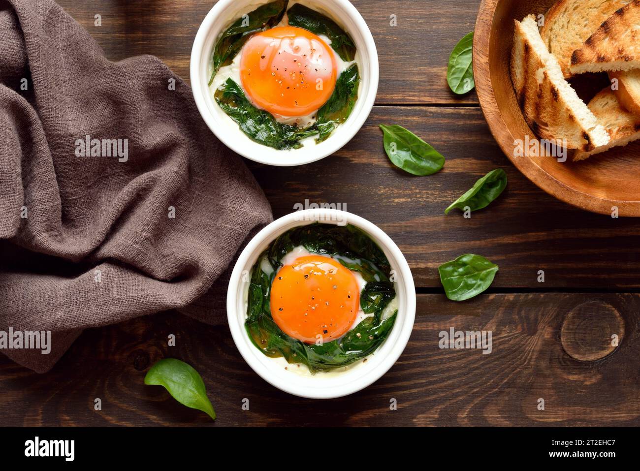 Eggs en cocotte (baked eggs) in ramekins with cream, сheese and spinach on wooden background. Top view, flat lay Stock Photo