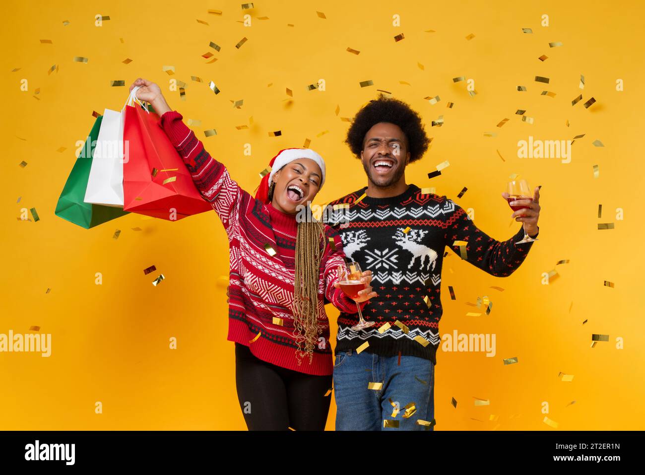 African Amercian couple wearing sweaters holding shoping bags and drinks celebrating Christmas in studio yellow color background Stock Photo
