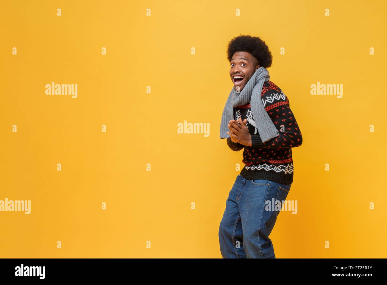 Happy young African-American man in Christmas sweater and scarf expressing surprise feeling in isolated yellow background with copy space studio shot Stock Photo