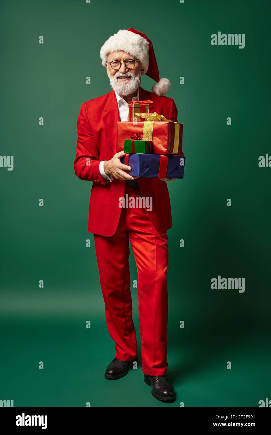elegant Santa in red stylish attire holding pile of presents on green backdrop, winter concept Stock Photo