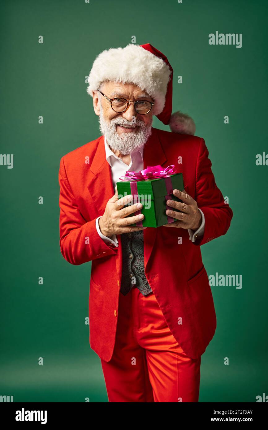 stylish Santa with beard and christmassy hat holding present in front of camera, winter concept Stock Photo