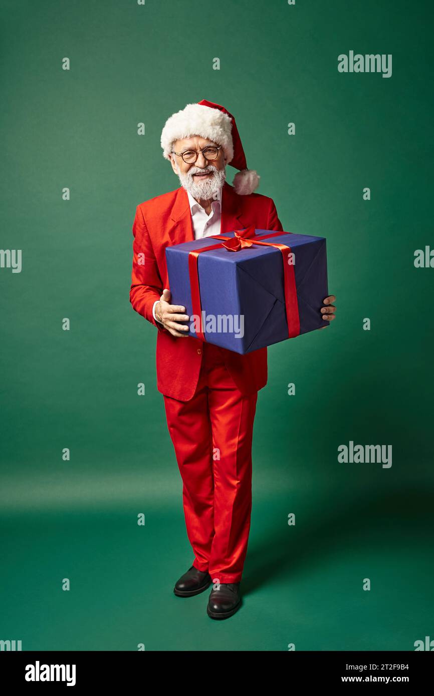elegant good looking Santa in christmassy hat holding huge blue present with ribbon, winter concept Stock Photo