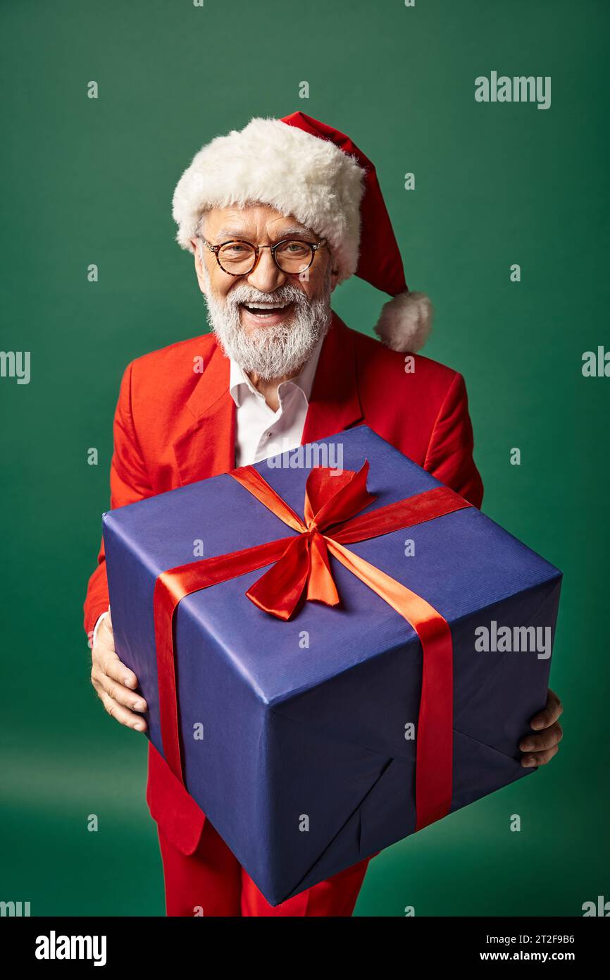 elegant jolly Santa in stylish red attire and hat showing big present at camera, winter concept Stock Photo