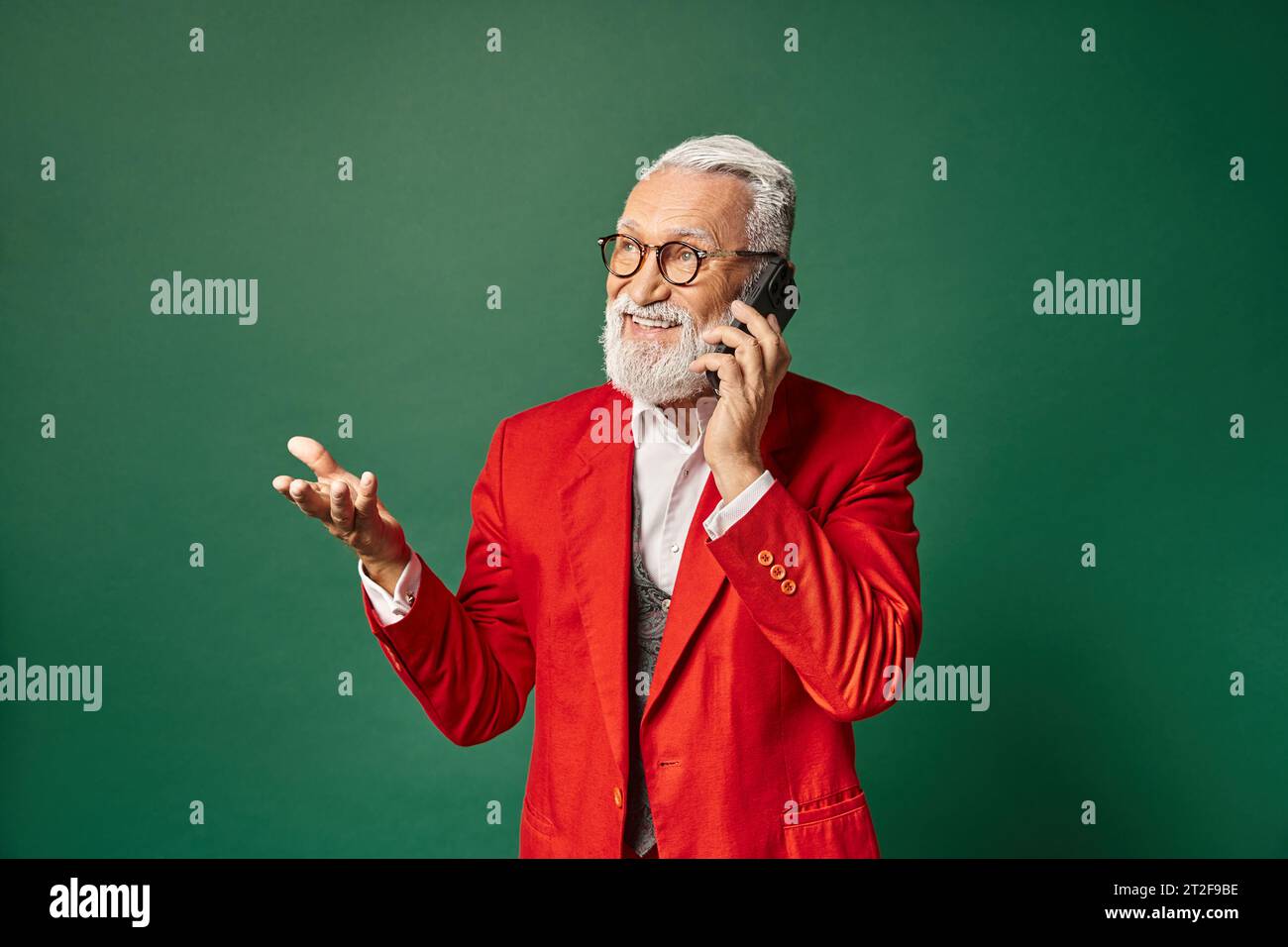 handsome Santa in stylish red attire gesturing and talking by phone cheerfully, winter concept Stock Photo