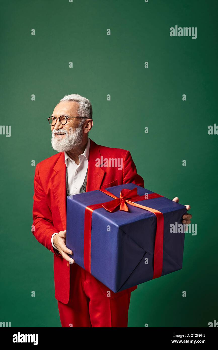 stylish classy Santa holding huge present on green backdrop and looking away, winter concept Stock Photo