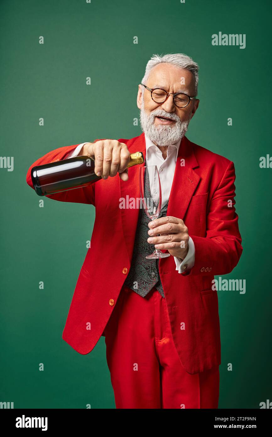 cheerful Santa with white beard pouring flute glass with champagne on green backdrop, winter concept Stock Photo