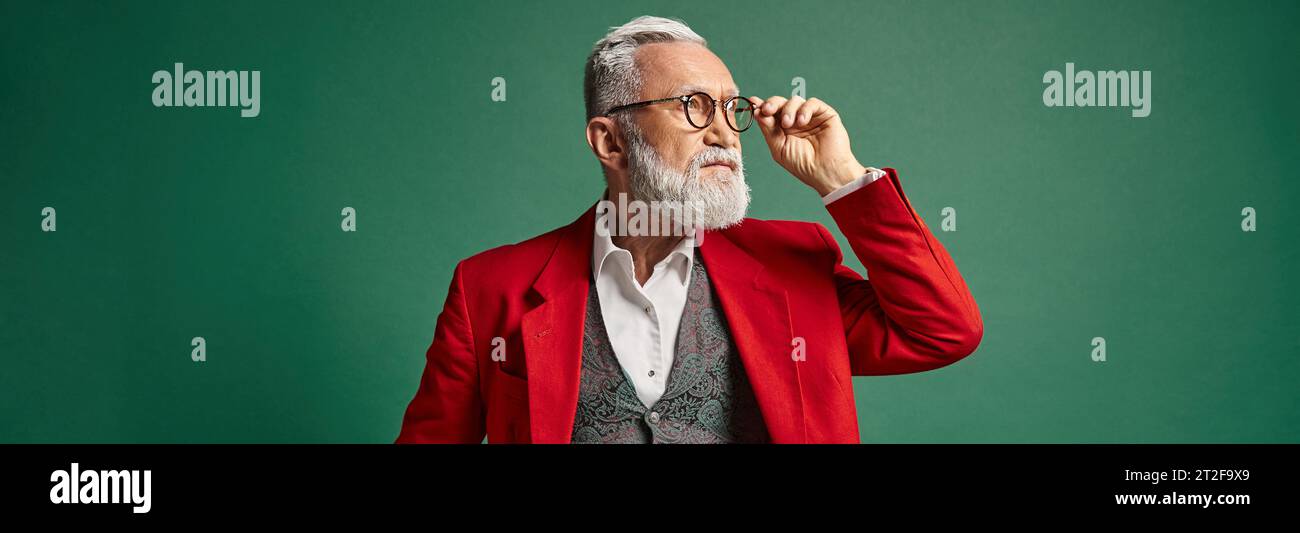 handsome Santa touching his glasses and looking away on green backdrop, winter concept, banner Stock Photo