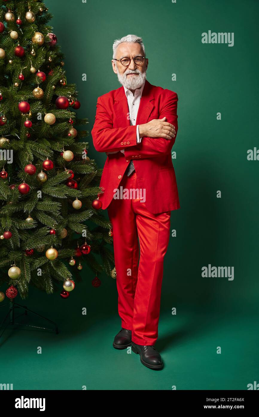 handsome Santa with white beard and glasses posing near fir tree with crossed arms, winter concept Stock Photo
