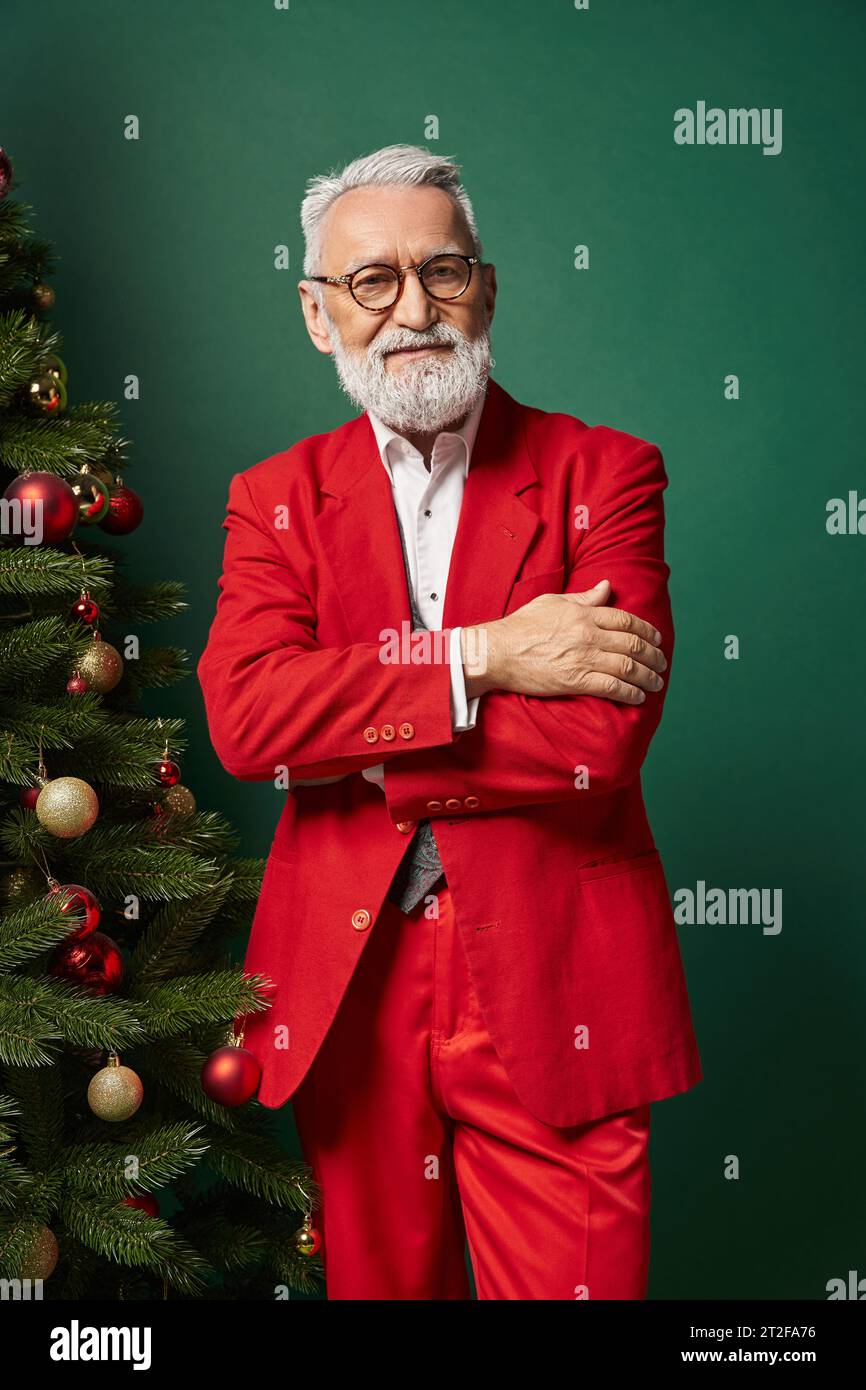 elegant Santa in red suit with glasses posing near Christmas tree with crossed arms, winter concept Stock Photo