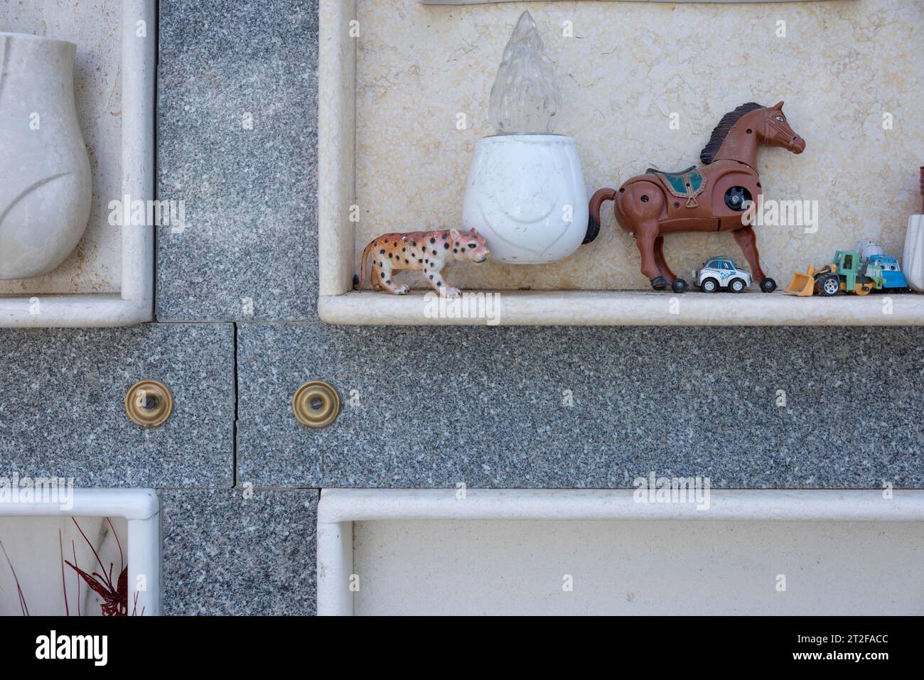 Toy, broken, mechanical horse with 3 legs, toy cars on an urn grave in a cemetery, Bari Sardo, Ogliastra, Sardinia, Italy Stock Photo