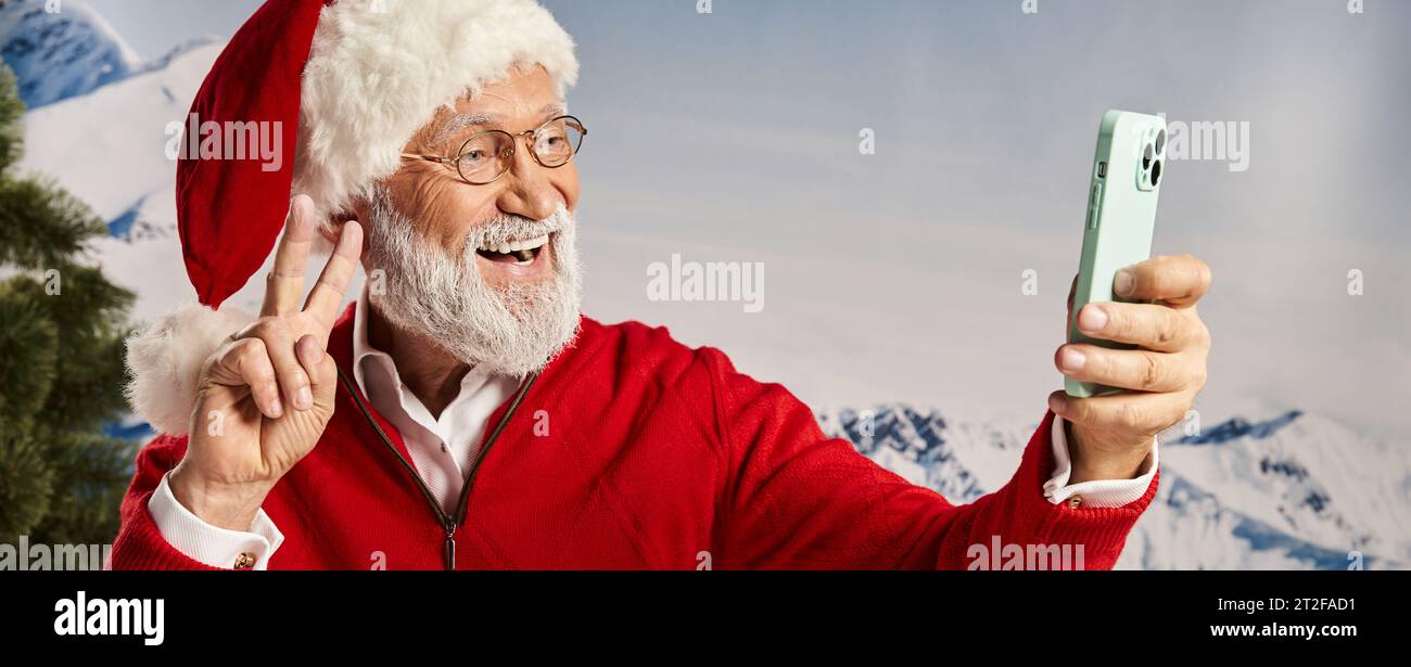 joyful man dressed as Santa wearing glasses and taking selfie showing peace gesture, winter, banner Stock Photo