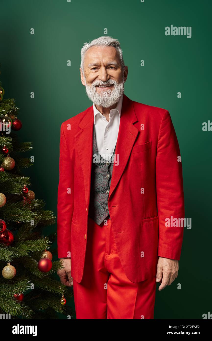 cheerful man in stylish Santa red costume posing near fir tree with baubles, Christmas concept Stock Photo