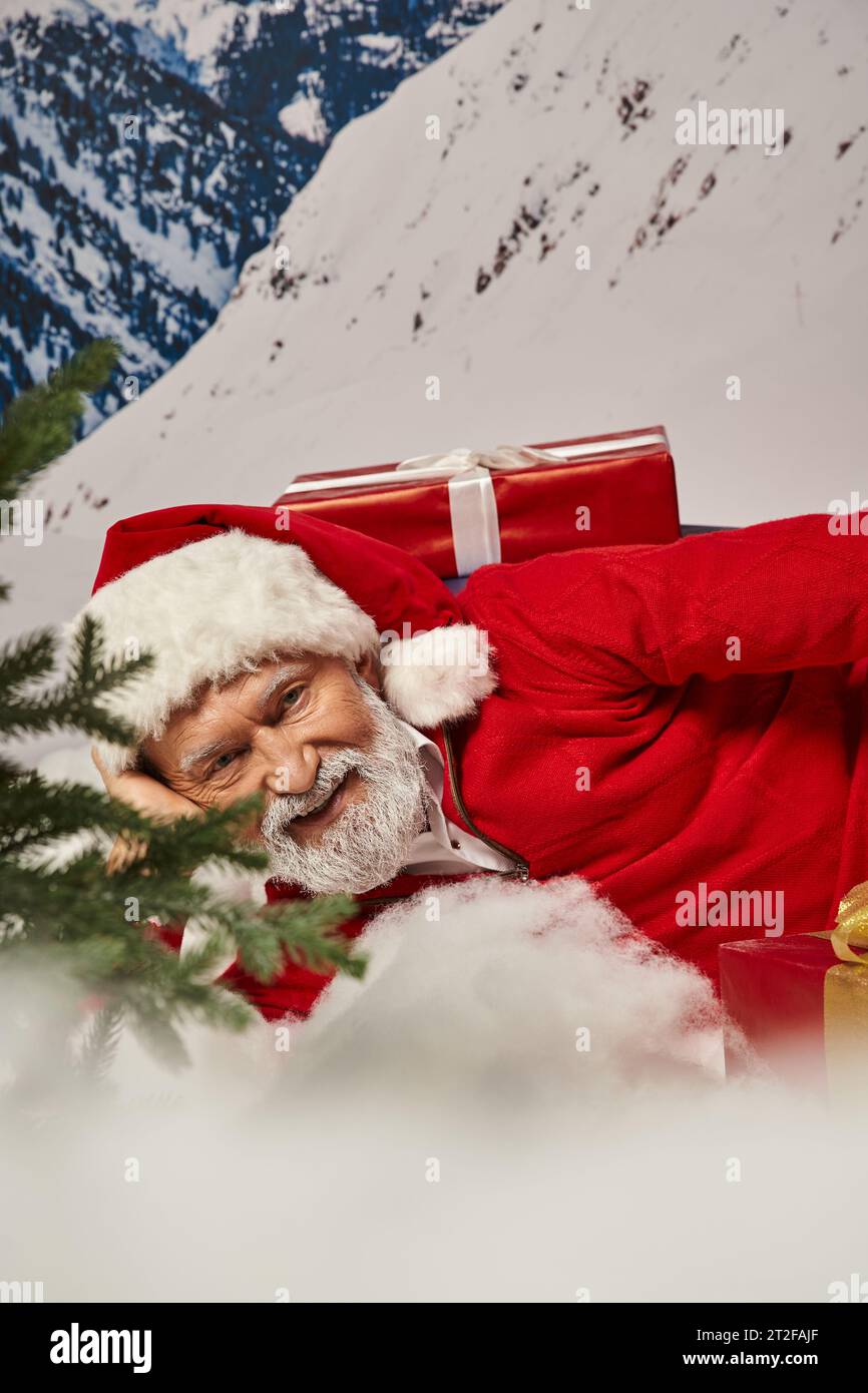 close up of joyous Santa in christmassy hat lying on snow surrounded by presents, winter concept Stock Photo