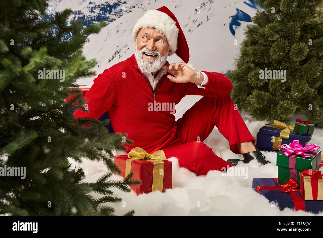 cheerful dreamy man in Santa costume posing on snow with presents looking away, Merry Christmas Stock Photo