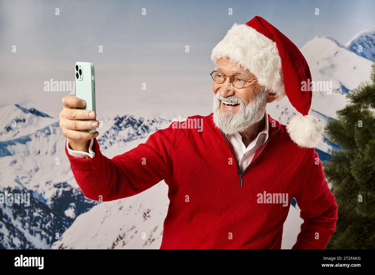 happy man dressed as Santa wearing glasses and taking selfie with mountain backdrop, winter concept Stock Photo