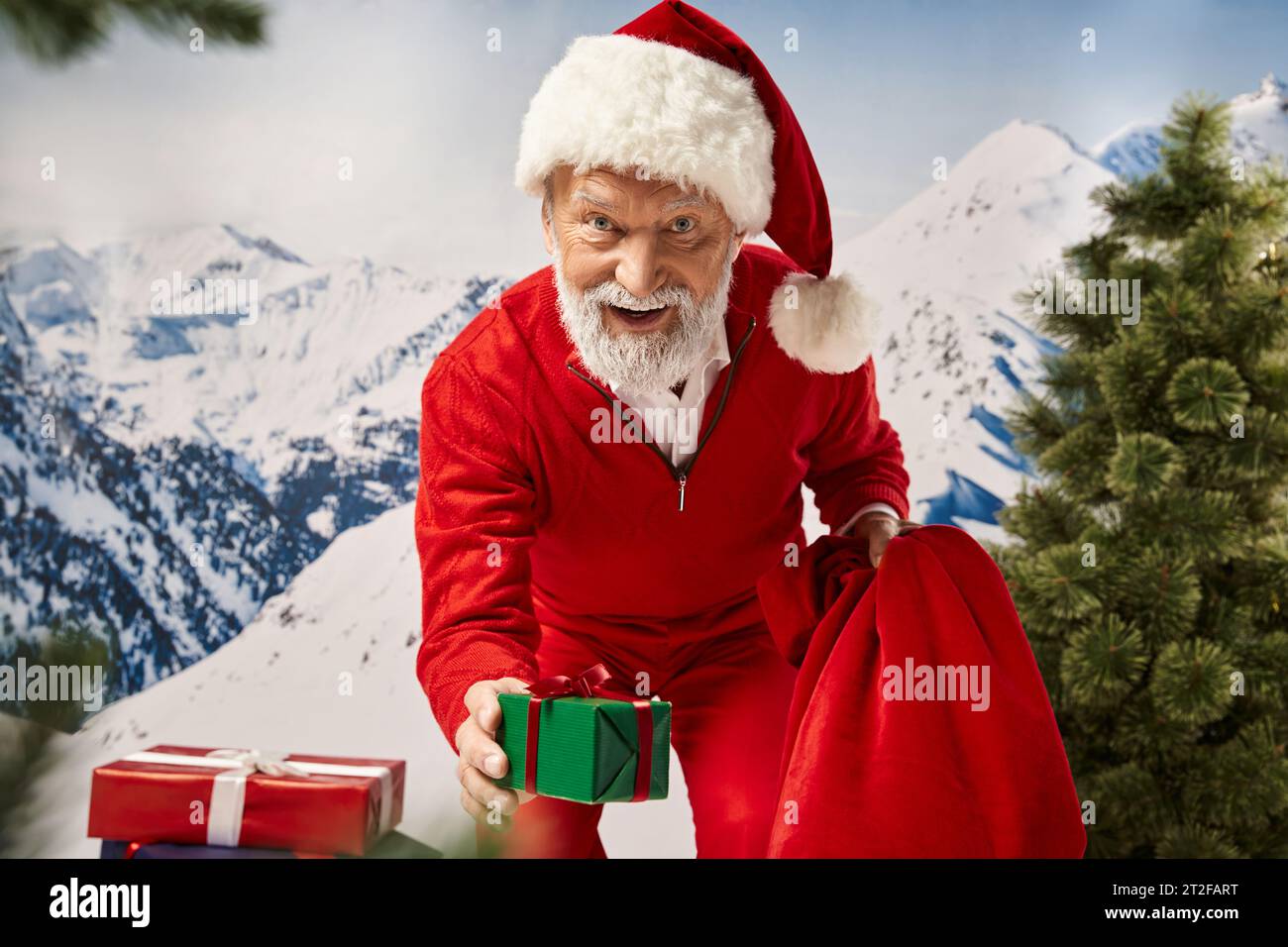 playful Santa surrounded by presents showing one at camera with slightly open mouth, winter concept Stock Photo