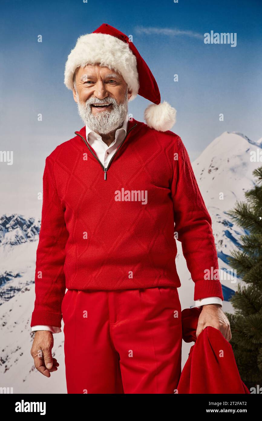 joyous Santa in warm red outfit posing with gift bag with mountain backdrop, winter concept Stock Photo