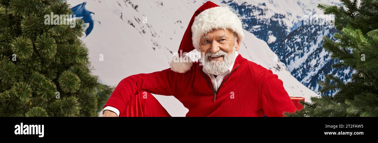 cheerful man dressed as Santa Claus in christmassy hat with snowy backdrop, winter concept, banner Stock Photo