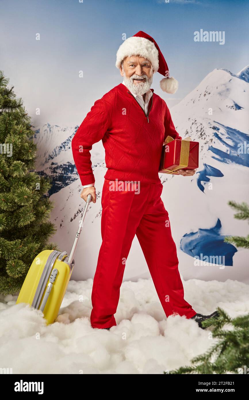 stylish Santa with white beard holding present in hand posing with yellow suitcase, winter concept Stock Photo