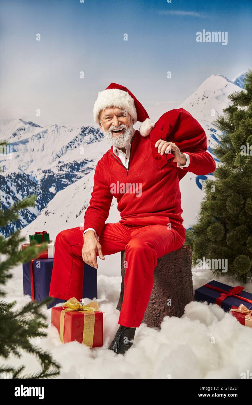 cheerful Santa sitting on tree stump surrounded by presents with snowy backdrop, winter concept Stock Photo