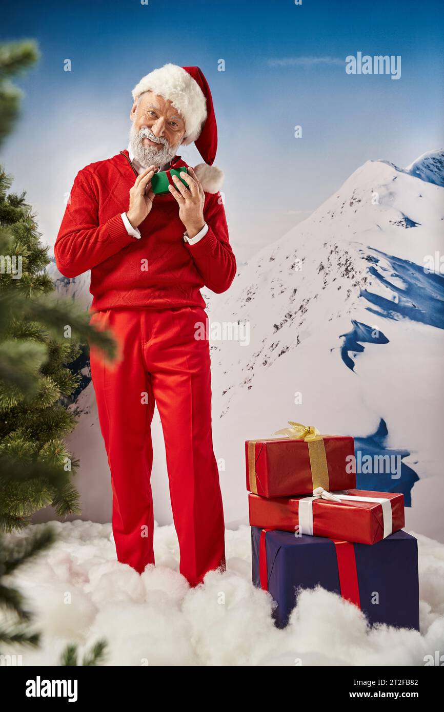 joyous Santa posing with presents and smiling at camera with mountain backdrop, winter concept Stock Photo