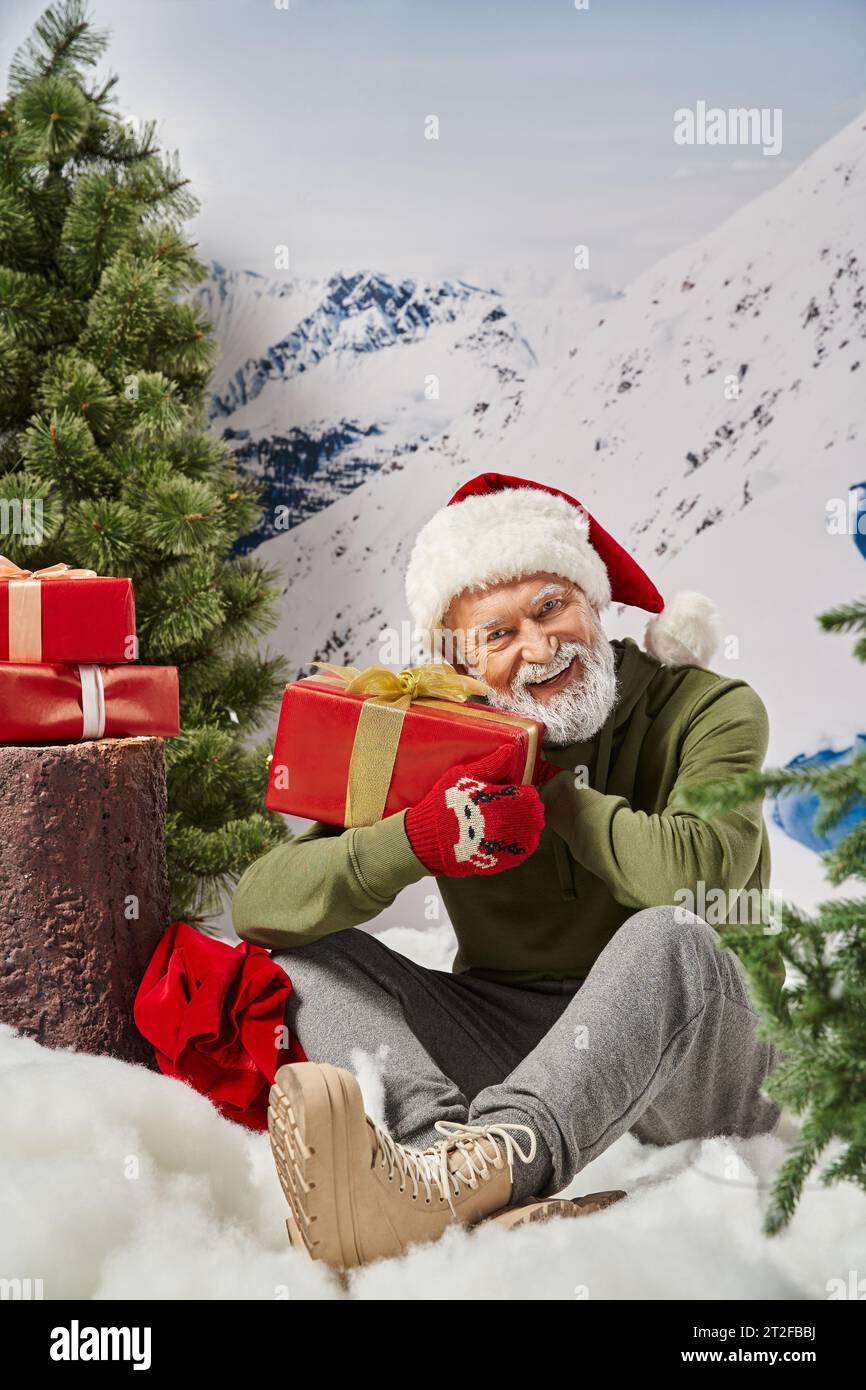 cheerful man dressed as Santa holding present and sitting on snow near tree stump, winter concept Stock Photo