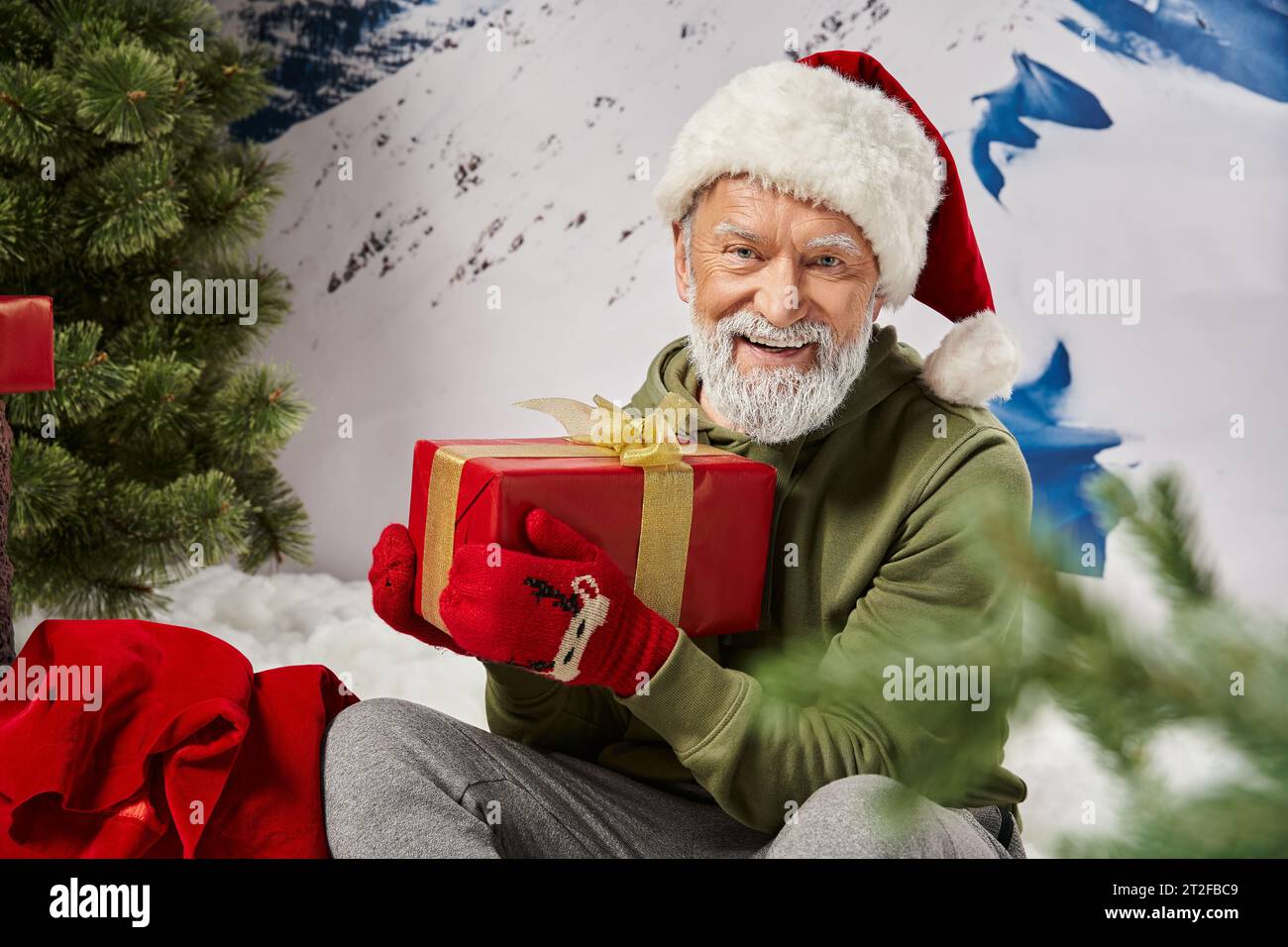 athletic Santa in red hat and mittens sitting with gift on snow near present bag, winter concept Stock Photo
