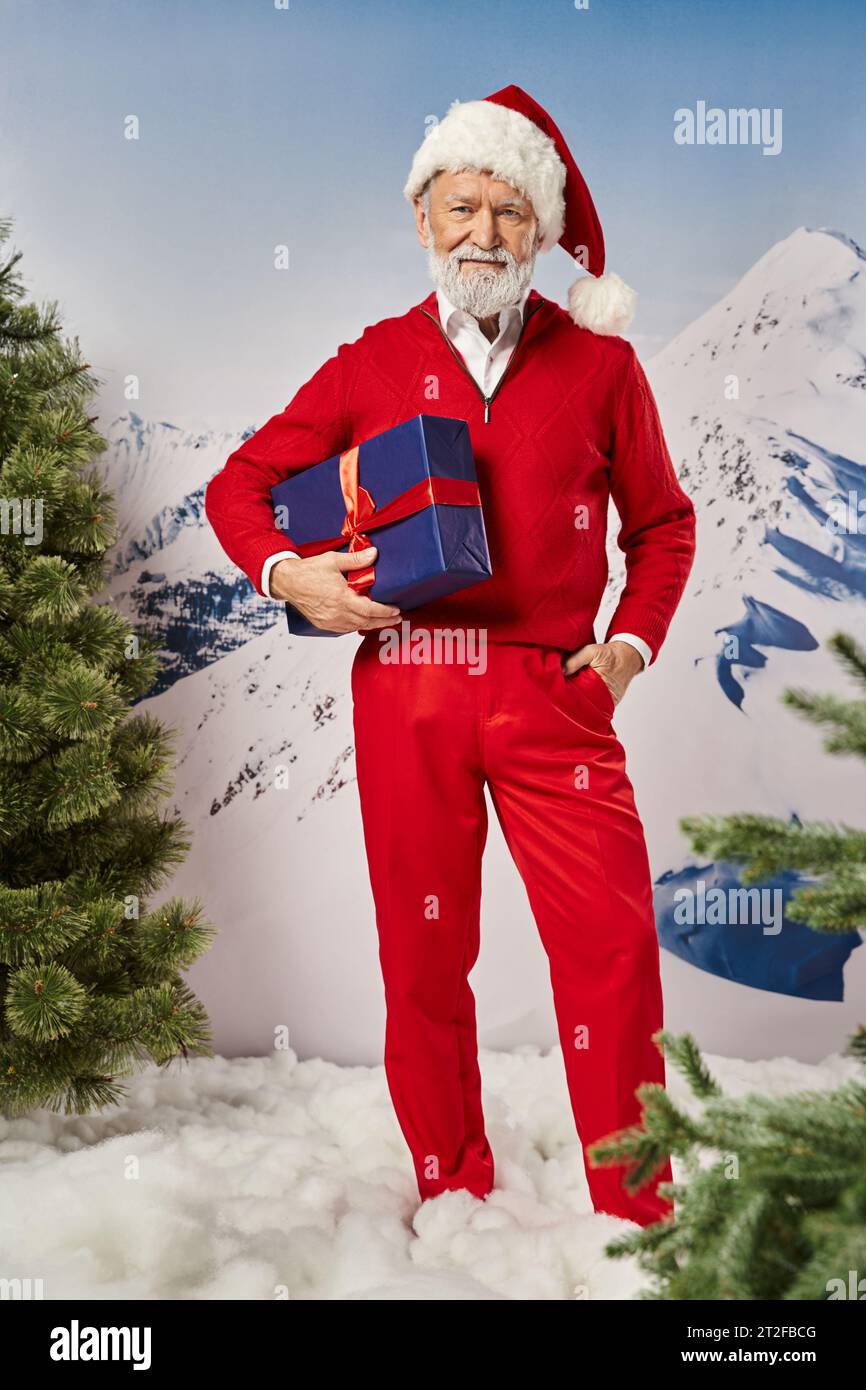 stylish Santa posing with present with one hand in pocket with mountain backdrop, winter concept Stock Photo