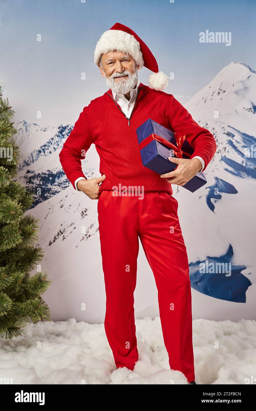 cheerful Santa holding present with one hand on hip with snowy mountain backdrop, winter concept Stock Photo