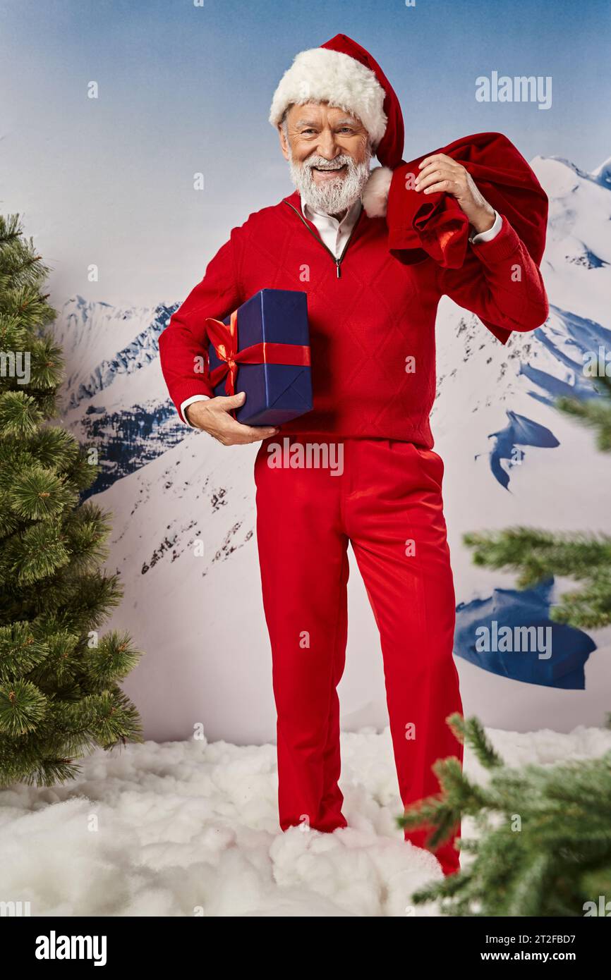 happy modern Santa with white beard in red outfit posing with gift and present bag, winter concept Stock Photo