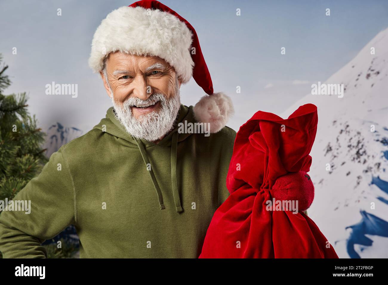 joyful athletic man in khaki hoodie wearing Santa hat and holding present bag, winter concept Stock Photo