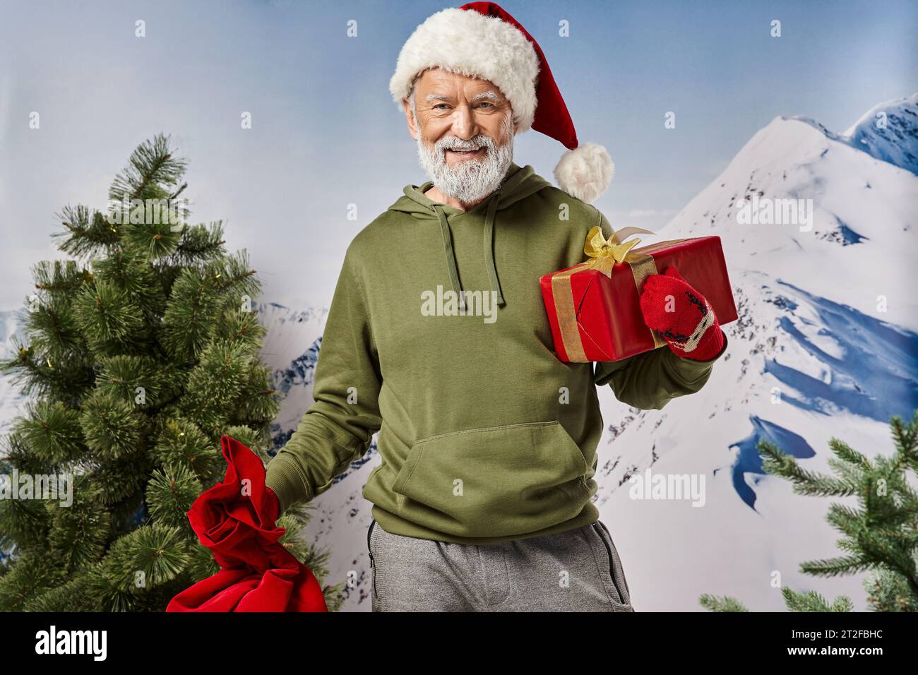 cheerful Santa in christmassy hat with present in hand with snowy mountain backdrop, winter concept Stock Photo