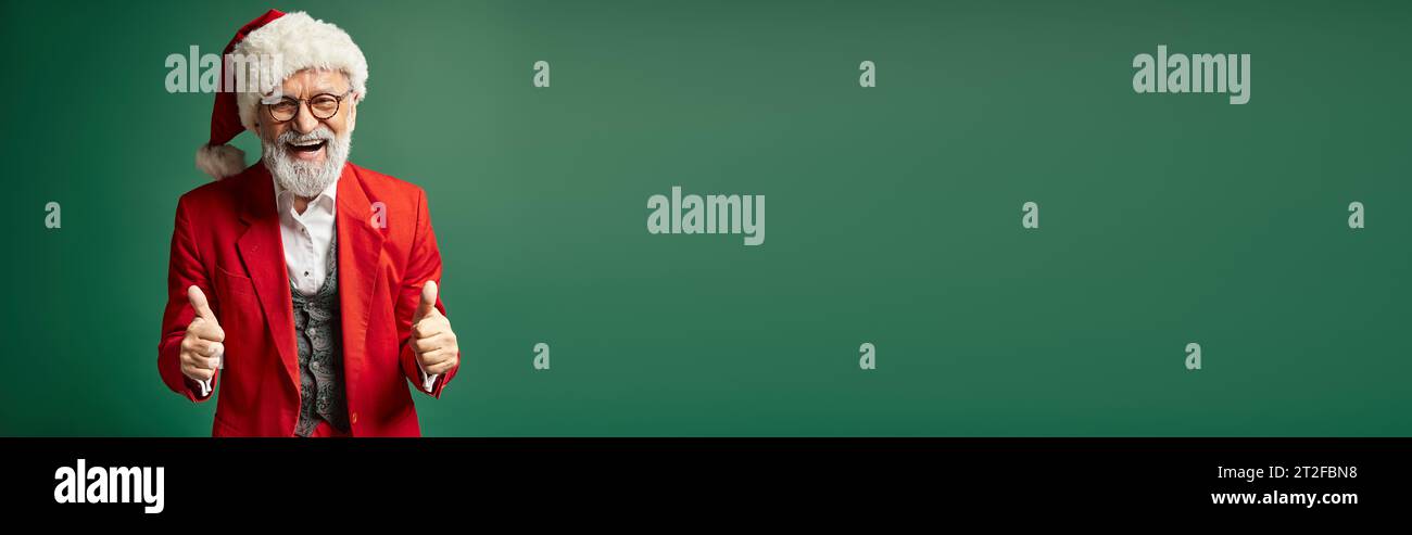 happy Santa on dark green backdrop smiling and showing thumbs up gesture, winter concept, banner Stock Photo