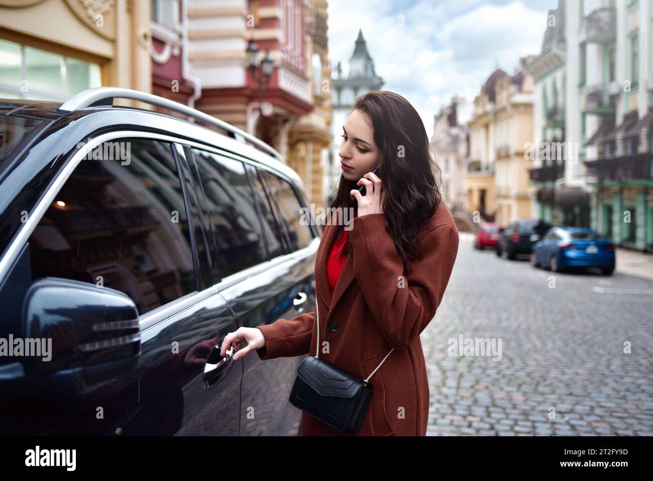 A Successful young woman with long hair in a red coat talks on the phone and gets into her car Stock Photo