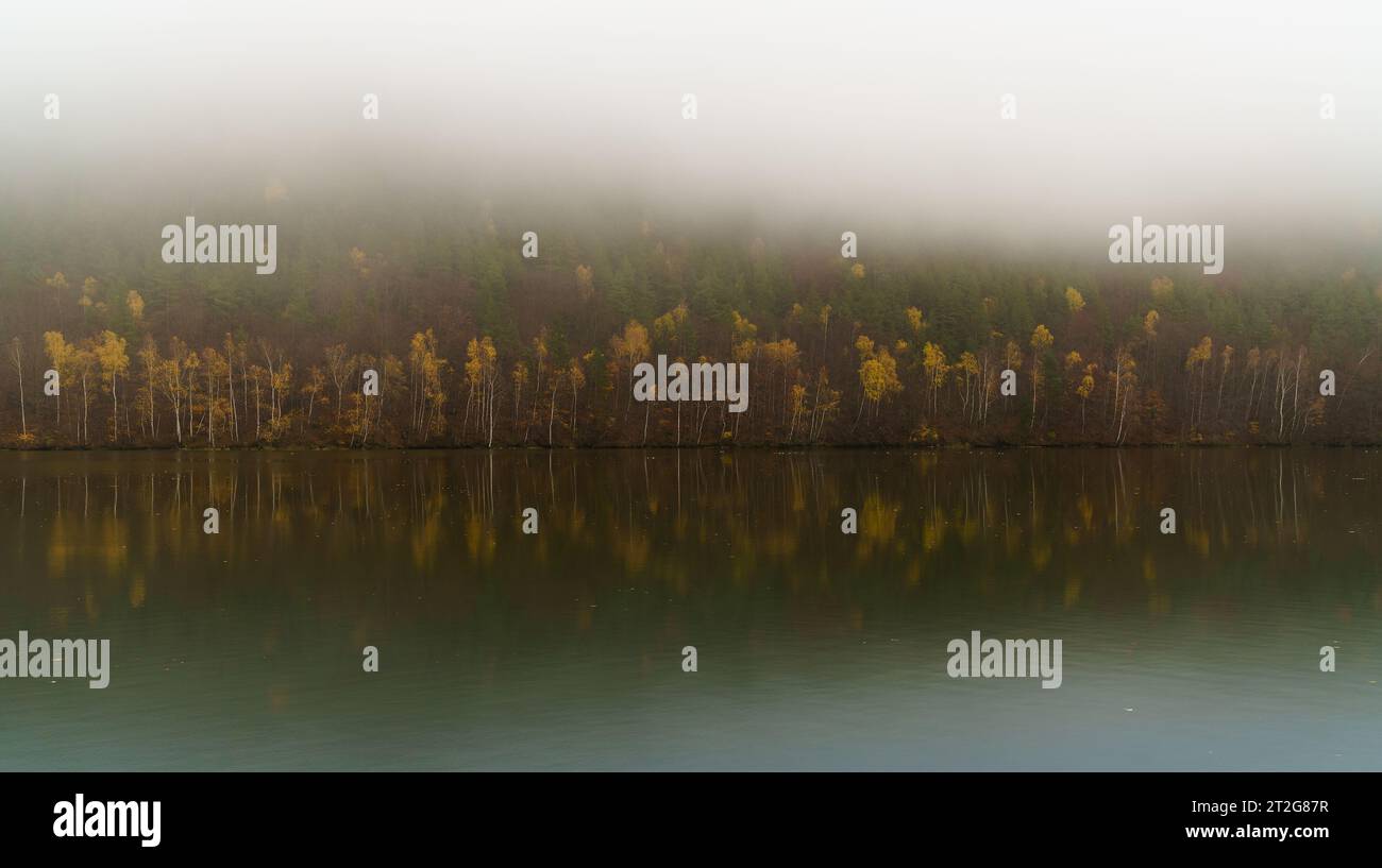 Wide Vag river calm autumnal waters with reflected yellow and red deciduous misty forest near Sutovo settlement, Slovak Republic. Moody weather and tr Stock Photo