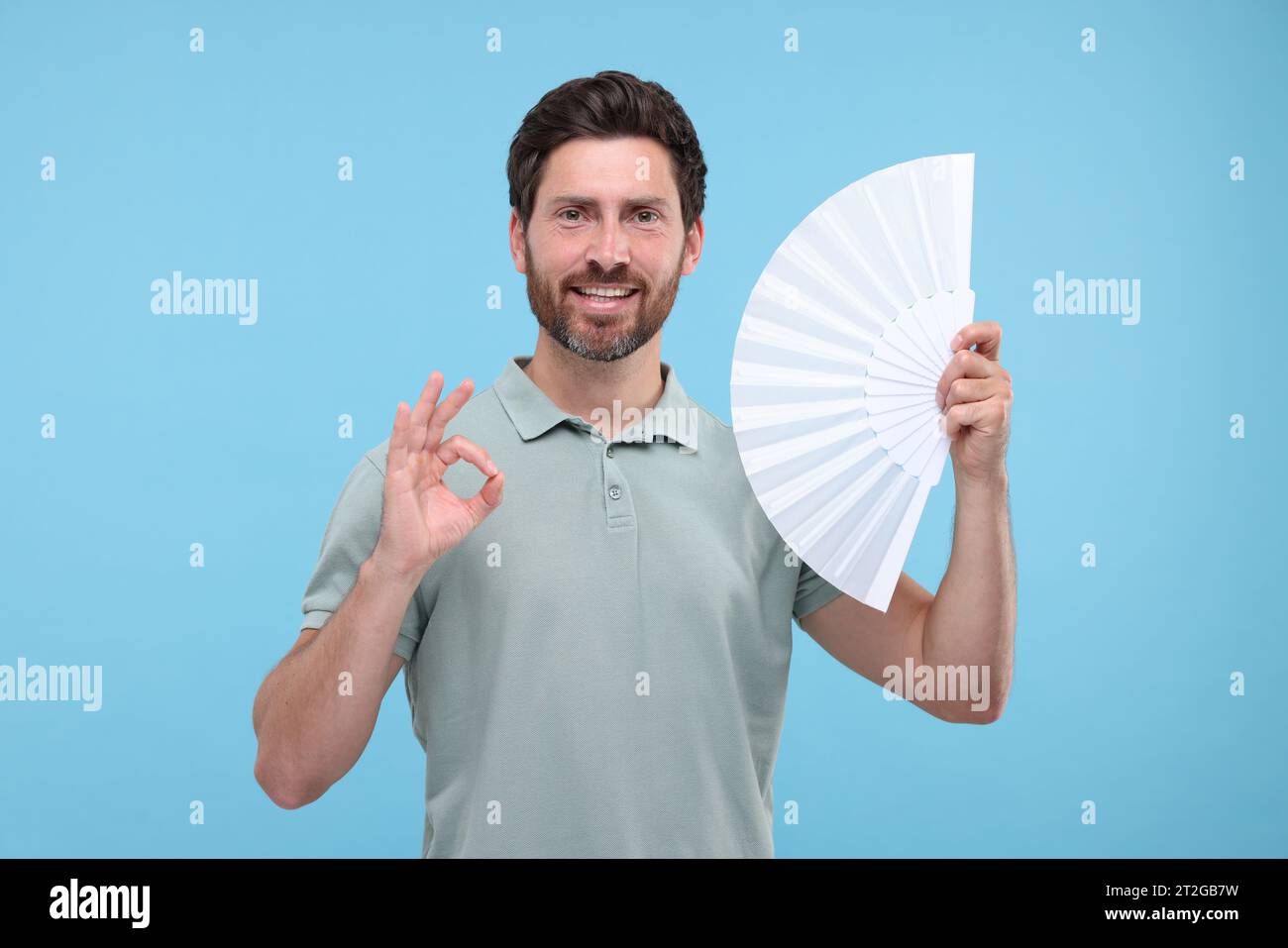 Happy man holding hand fan and showing ok gesture on light blue ...