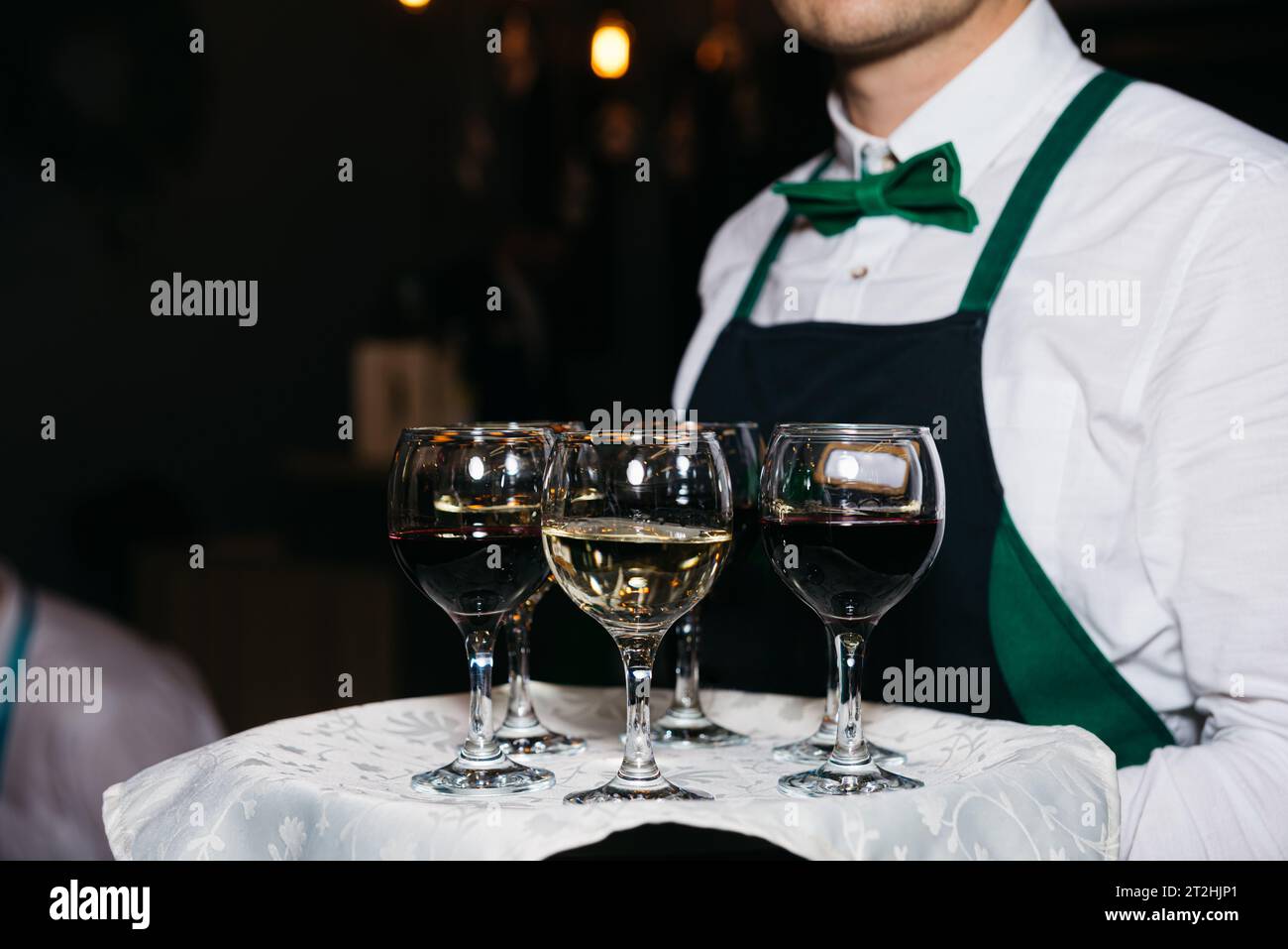 The waiter hold tray with several glasses of red and white wine. The employee wears a black apron, white shirt and green bowtie. Stock Photo