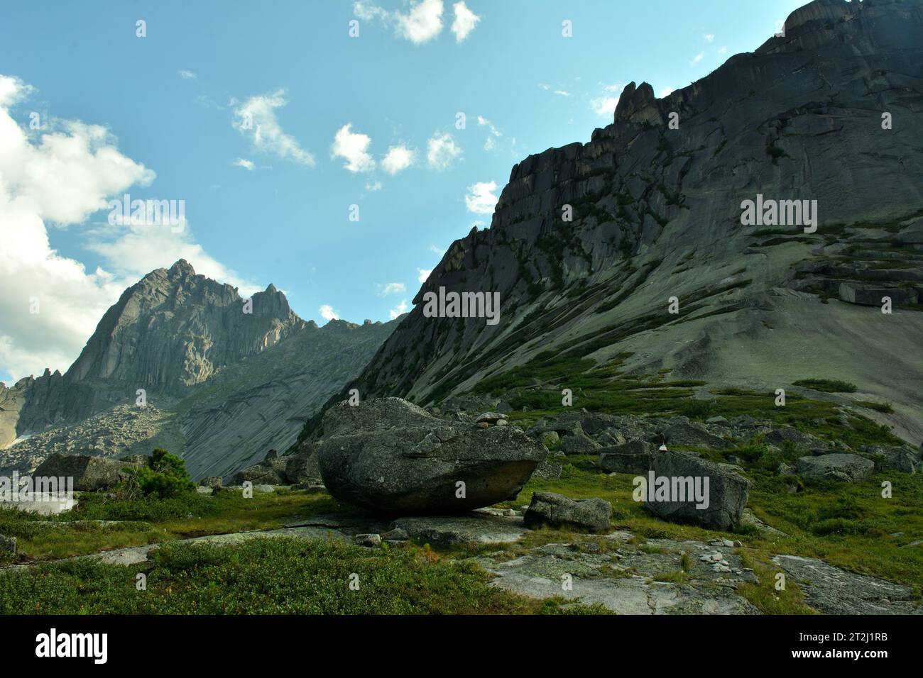 A scattering of large stones in a wide clearing with low grass at the foot of a high sheer cliff. Natural park Ergaki, Krasnoyarsk region, Siberia, Ru Stock Photo