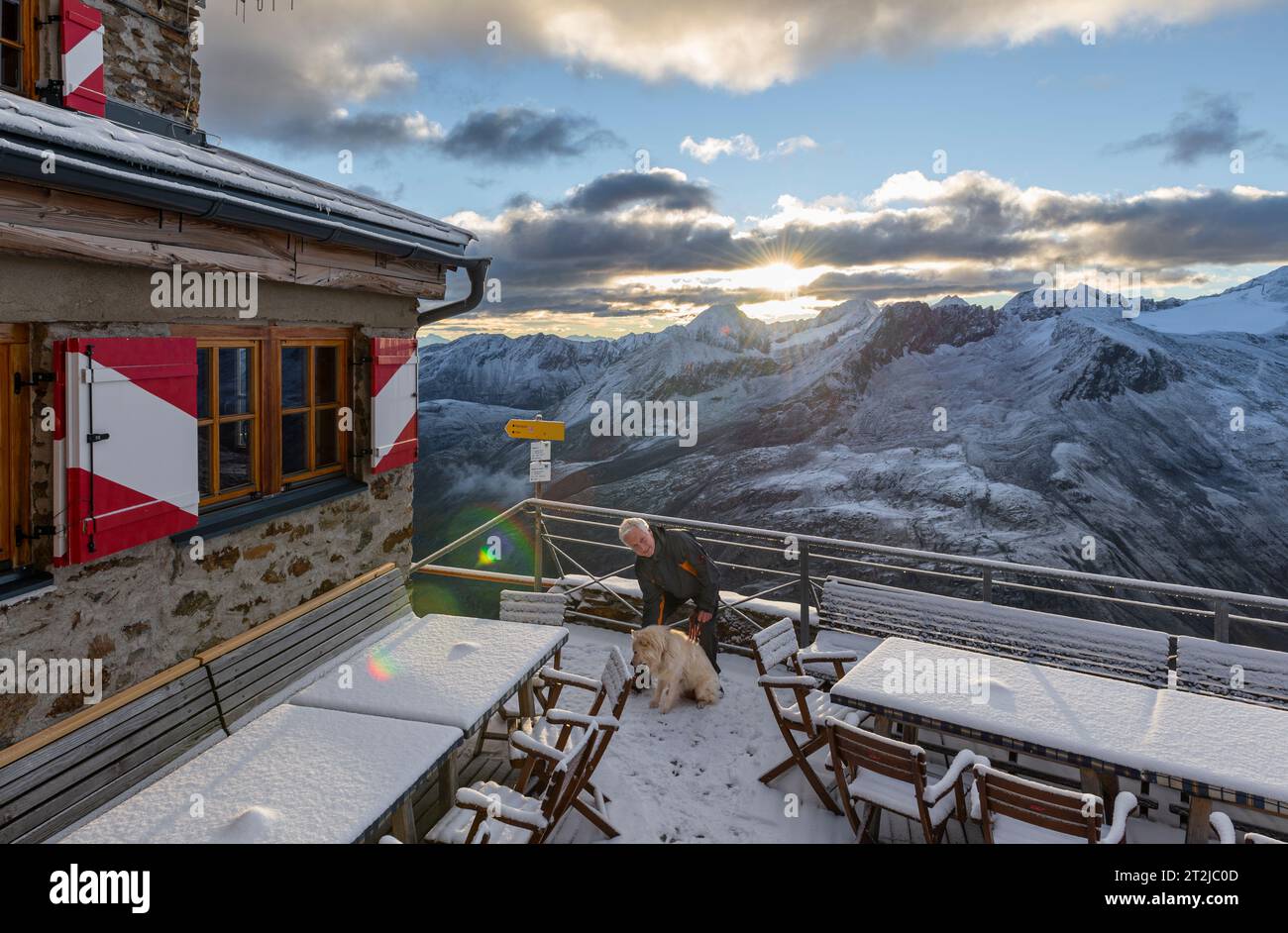 Man with dog in front of sunrise above Ramolhaus mountain hut with view of the cloud-covered main ridge of the Ötztal and Stubai Alps Stock Photo