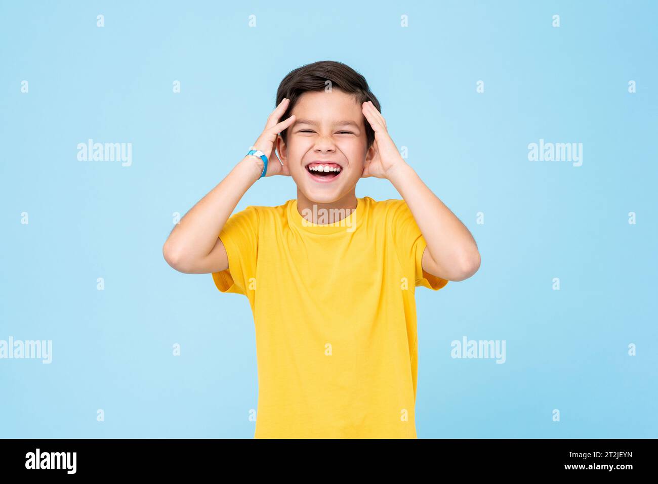 Excited mixed race boy in yellow t shirt touching head and laughing out loud against blue background Stock Photo
