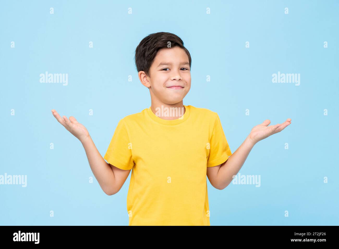 Cute bewildered mixed race boy in yellow t shirt looking at camera with smile and shrugging shoulders against blue background Stock Photo