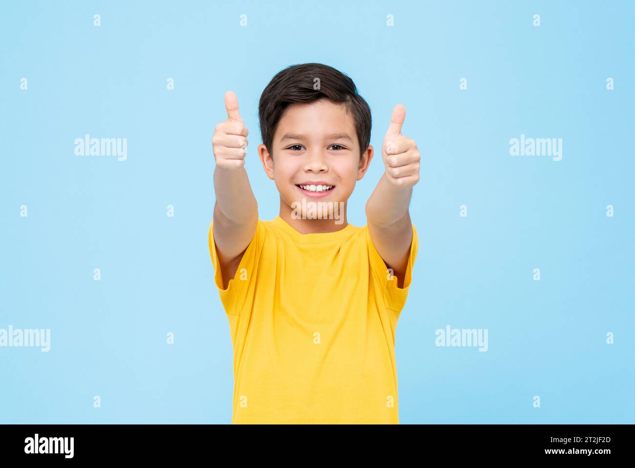 Happy mixed race boy in yellow t shirt smiling and looking at camera while showing thumbs up gesture in approval against blue background Stock Photo