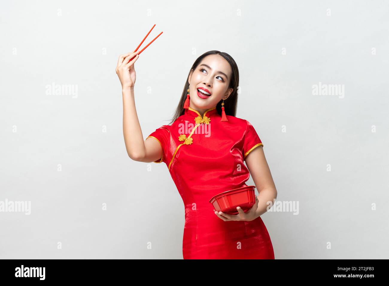 Cheerful Asian female in traditional red dress raising arm with chopsticks and looking up with smile against gray background Stock Photo