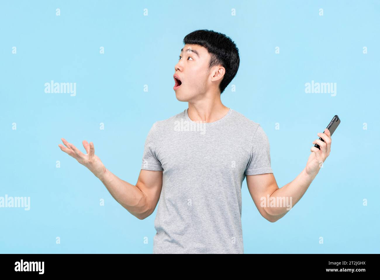 Astonished Asian man with mobile phone in hands looking up while standing on light blue isolated background in studio Stock Photo