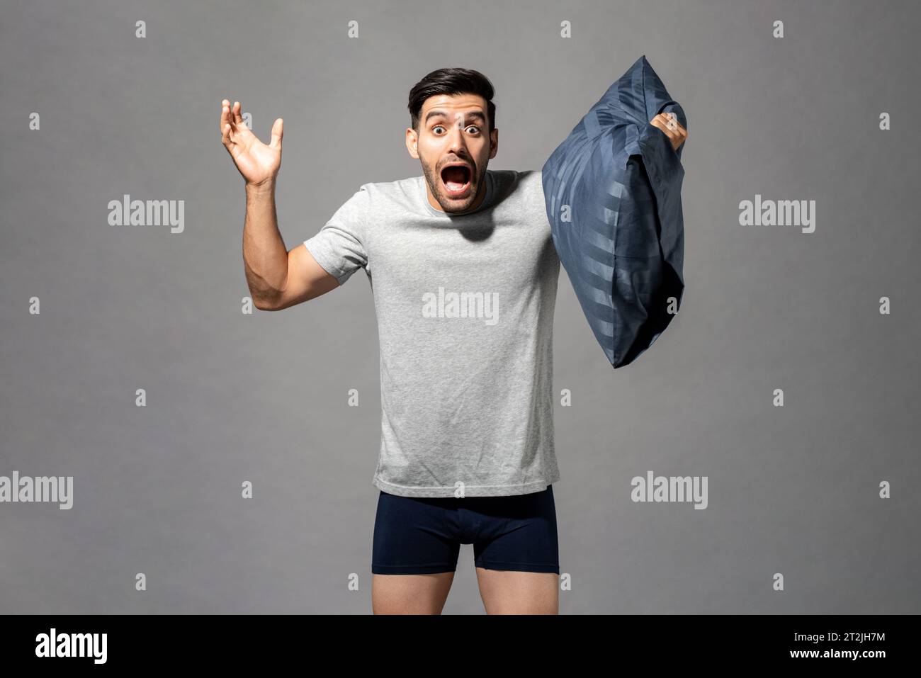 Astonished Caucasian man in nightwear with pillow in hand screaming with raised arms on gray background Stock Photo