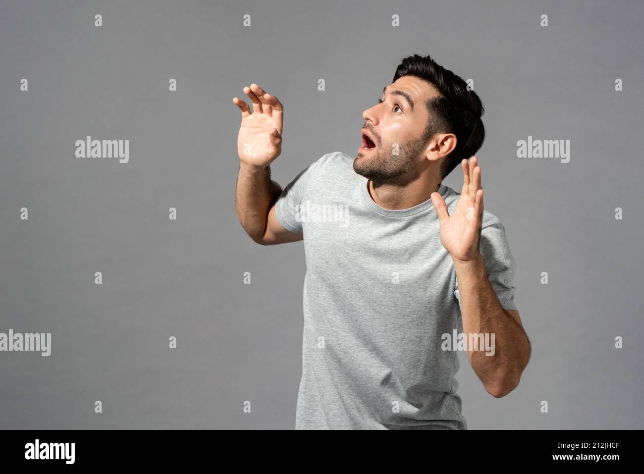 Frightened bearded Caucasian man in t shirt looking up with raised hands while standing on gray background in light studio Stock Photo