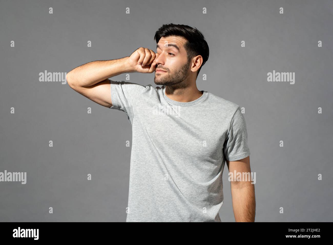 Sleepy bearded Caucasian man in casual t shirt rubbing eye while standing on gray isolated background in the morning Stock Photo
