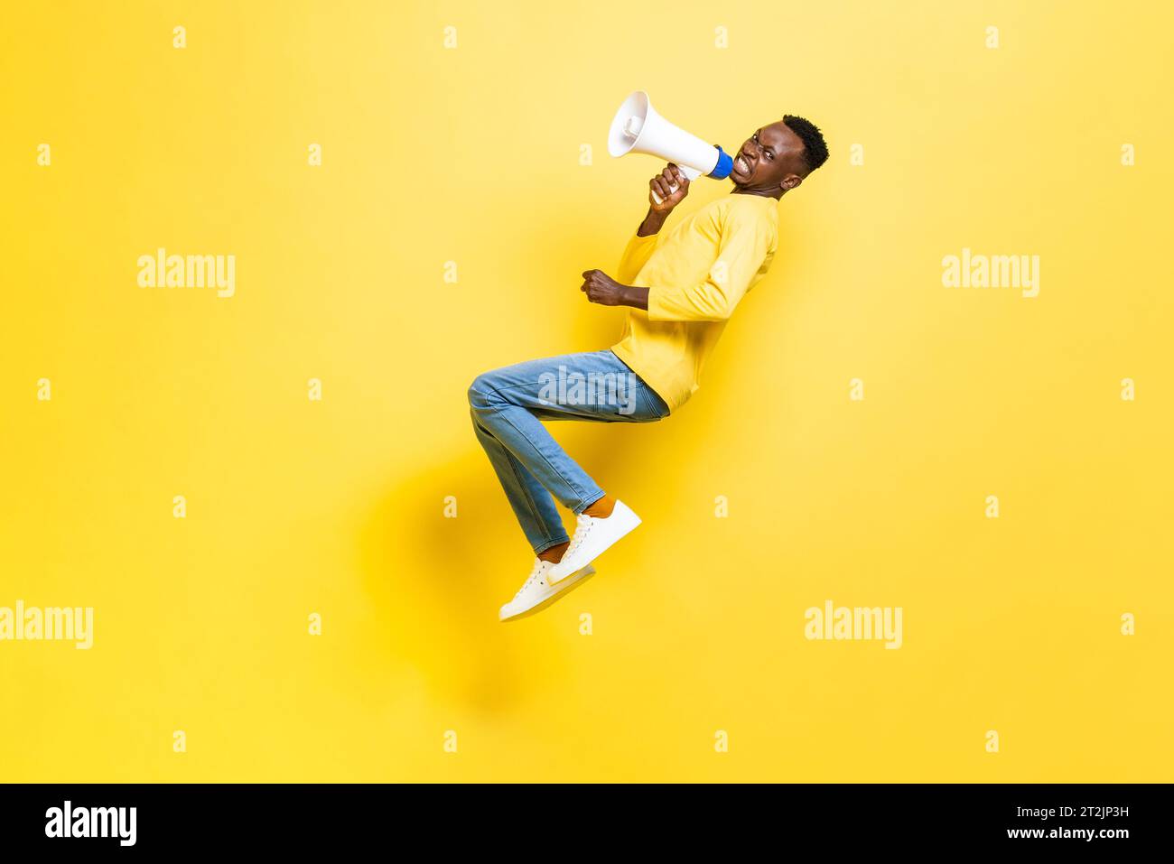 Full body side view of African angry man screaming in megaphone and looking at camera while levitating on yellow background Stock Photo