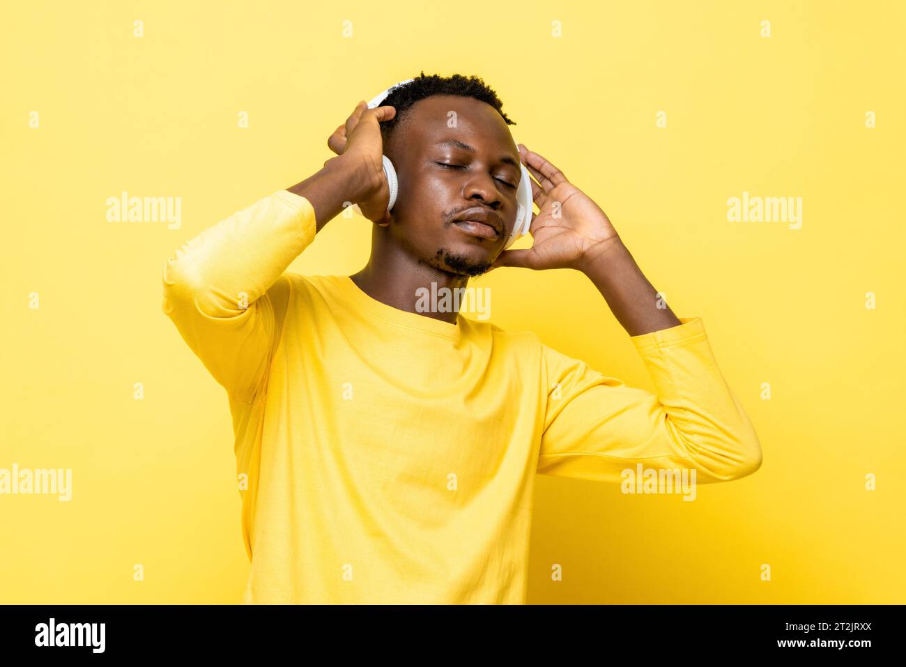 Young African man listening to music with eyes closed while holding headphones against yellow studio background Stock Photo