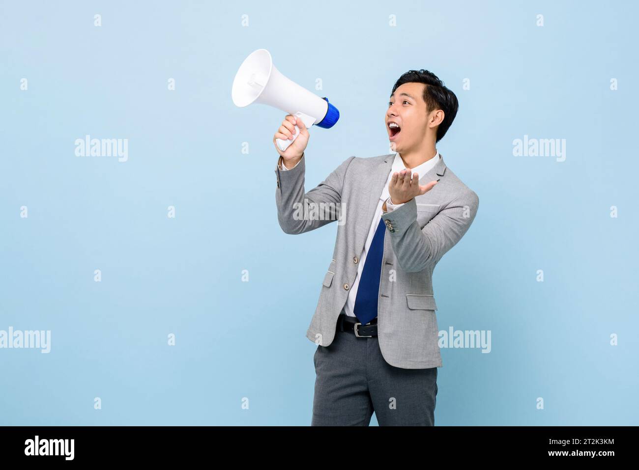 Young handsome Asian man in formal business suit holding megaphone and yelling in isolated light blue color studio background Stock Photo
